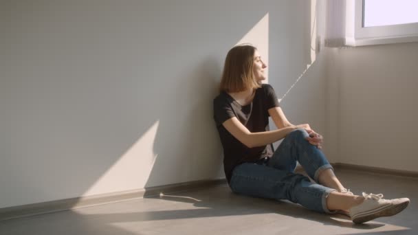 Крупный план портрета молодой веселой кавказки, сидящей на полу у окна и счастливо улыбающейся в уютной солнечной квартире — стоковое видео