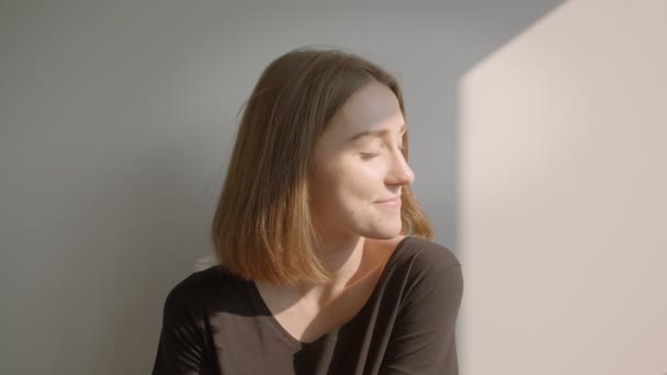 Gros plan portrait de jeune femme caucasienne détendue assise sur le sol près de la fenêtre souriant joyeusement à l'intérieur dans le confortable appartement ensoleillé — Video