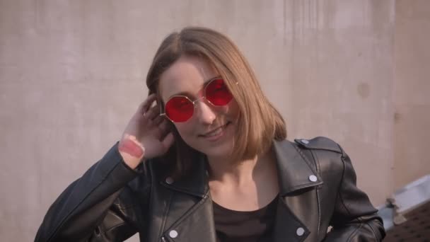 Gros plan portrait de jeune fille caucasienne mignonne dans une veste en cuir et lunettes de soleil hipster rouge souriant joyeusement regarder la caméra en plein air dans la ville urbaine — Video