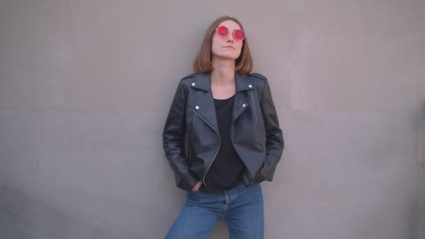 Primer plano retrato de joven linda chica caucásica en una chaqueta de cuero y gafas de sol rojas posando frente a la cámara al aire libre en la ciudad urbana — Vídeos de Stock