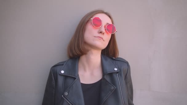 穿着皮夹克和红色太阳镜的年轻酷白种女孩的特写肖像，在城市户外的摄像机前摆姿势 — 图库视频影像