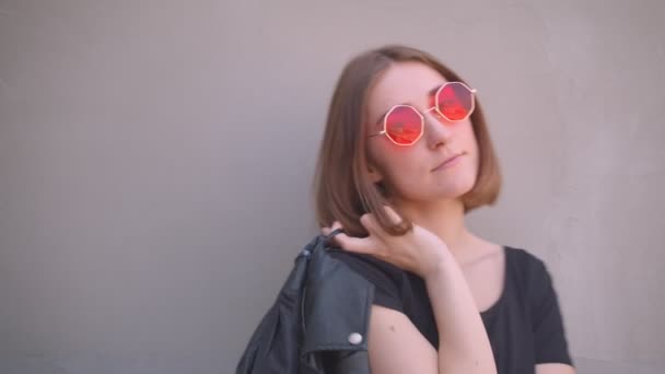 Retrato de cerca de una joven caucásica fresca en gafas de sol rojas con chaqueta de cuero sobre el hombro sonriendo posando frente a la cámara al aire libre en la ciudad urbana — Vídeos de Stock