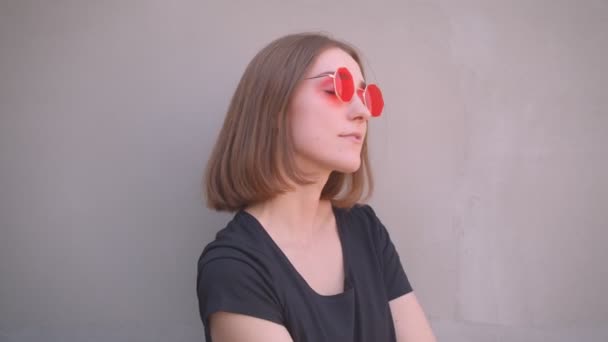 Retrato de cerca de una joven caucásica fresca en gafas de sol rojas con chaqueta de cuero sobre el hombro sonriendo mirando a la cámara al aire libre en la ciudad urbana — Vídeo de stock