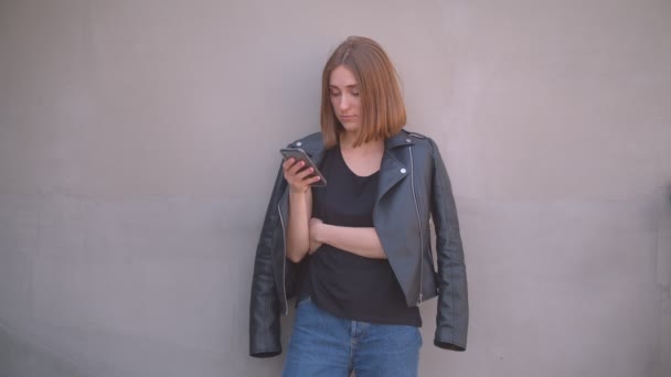 Zbliżenie portret młodego cute kaukaski dziewczyna w skórzanej kurtce za pomocą telefonu przed kamerą na zewnątrz w miejskim mieście — Wideo stockowe