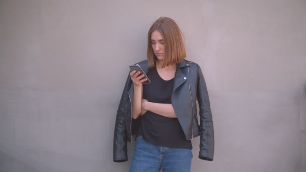 Primer plano retrato de joven linda chica caucásica en una chaqueta de cuero usando el teléfono mirando a la cámara al aire libre en la ciudad urbana — Vídeo de stock