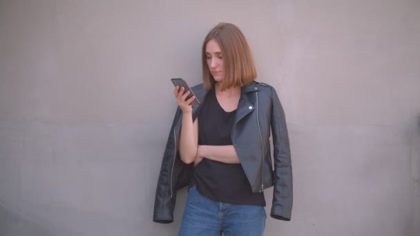 Крупный план портрет юной милой белой девушки в кожаной куртке с помощью телефона, счастливо улыбающейся, глядя в камеру на открытом воздухе в городе — стоковое видео