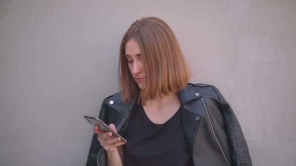 Closeup portrait of young pretty caucasian girl in a leather jacket using the phone looking at camera outdoors in the urban city — Stock Video