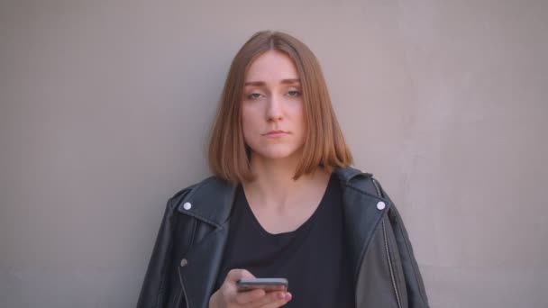 Zbliżenie portret młodego całkiem kaukaski dziewczyna w skórzanej kurtce za pomocą telefonu patrząc na kamerę na zewnątrz w miejskim mieście — Wideo stockowe