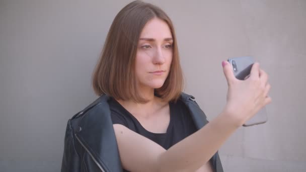 Closeup portrait of young pretty caucasian girl in a leather jacket taking selfies on the phone outdoors in the urban city — Stock Video