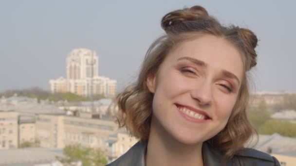 Closeup shoot of young pretty caucasian female with hair buns looking at camera with urban landscape on the background — Stock Video
