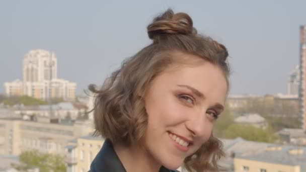 Zbliżenie strzelać z młody ładne kaukaski żeński z włosy bułeczki uśmiechnięty i szczęśliwie patrząc w aparat fotograficzny rezygnować miejski krajobraz od tło — Wideo stockowe
