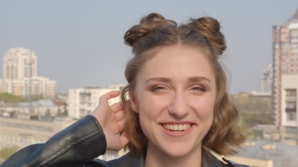 Closeup shoot of young pretty caucasian female with hair buns smiling and happily posing in front of the camera with urban landscape on the background — Stock Video