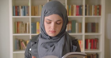 Türbanlı genç çekici Müslüman kız öğrencinin kapalı alanda üniversite kütüphanesinde kameraya bakan bir kitap okumasının yakın çekim portresi