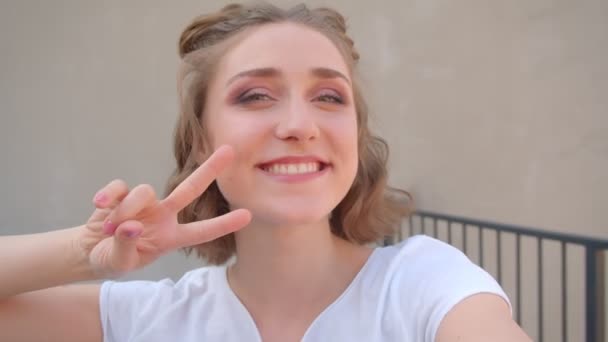 Primer plano de disparar de joven linda hembra caucásica con bollos de pelo tomando selfies en el teléfono mostrando dos dedos sonriendo felizmente mirando a la cámara con la ciudad urbana en el fondo — Vídeo de stock