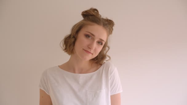 Closeup shoot of young cute caucasian female with hair buns smiling joyfully looking at camera in the white room indoors — Stock Video