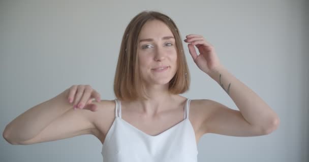 Съемка крупным планом молодой довольно белой женщины с кольцами волос, позирующими перед камерой в белом помещении — стоковое видео