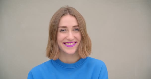 Крупним планом портрет молодої короткошерстої жінки з фіолетовою помадою посміхаючись і сміючись дивлячись на камеру з вуличною стіною на фоні — стокове відео