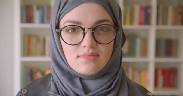 Retrato de primer plano de una joven atractiva estudiante musulmana en hijab y gafas mirando a la cámara en la biblioteca universitaria en el interior — Vídeo de stock