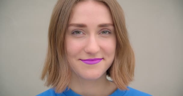 Closeup portrait of young short haired brunette female with purple lipstick smiling cheerfully posing in front of the camera with street wall on the background — Stock Video