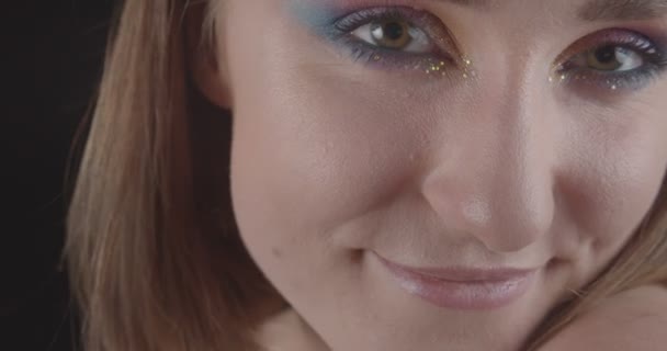Närbild porträtt av unga charmiga kaukasiska korthårig kvinnlig ansikte med söt glitter makeup titta på kamera leende med bakgrund isolerade på svart — Stockvideo