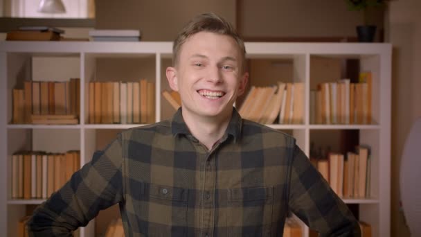 Съемки крупным планом молодого привлекательного кавказского студента, улыбающегося и счастливо смеющегося, глядя в камеру в библиотеке колледжа — стоковое видео