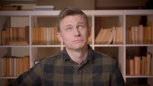 Primer plano de disparar de joven atractivo caucásico estudiante masculino mostrando la lengua y haciendo expresiones faciales divertidas mirando a la cámara en la biblioteca de la universidad — Vídeo de stock