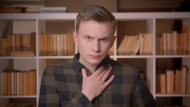 Gros plan shoot of young attractive caucasian male student having a sick throat looking at camera in the college library — Video