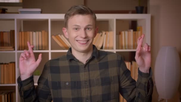 Gros plan shoot of young attractive caucasien mâle étudiant ayant ses doigts croisés être plein d'espoir en regardant la caméra dans la bibliothèque du collège — Video