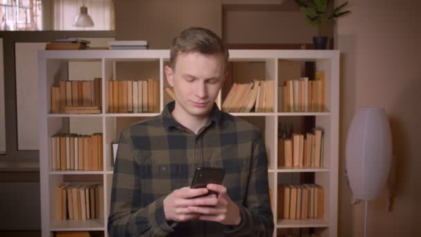 Närbild shoot av unga attraktiva kaukasiska manliga studenten använda telefonen i kollegiet biblioteket — Stockvideo