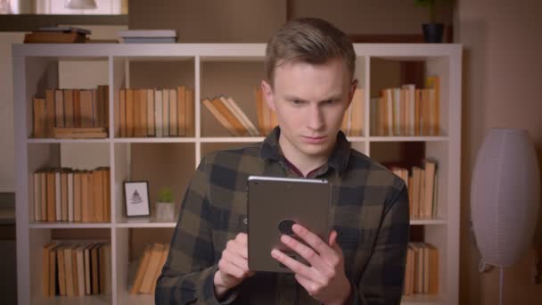 Gros plan shoot of young attractive caucasian male student browsing on the tablet looking at camera in the college library — Video