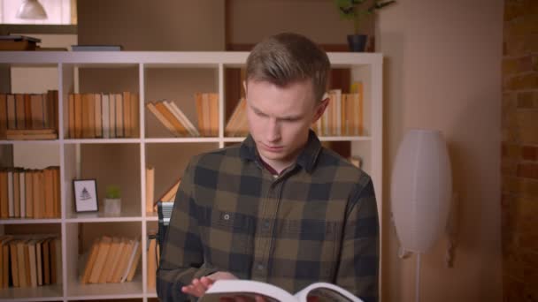 Sesión de primer plano de joven estudiante caucásico atractivo leyendo un libro mirando la cámara en la biblioteca de la universidad — Vídeos de Stock