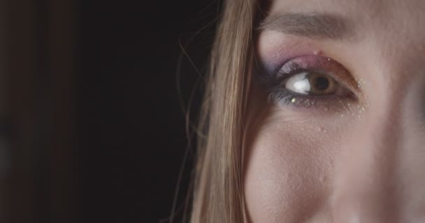 Primo piano mezza faccia ritratto di giovane femmina caucasica con occhi con trucco glitter guardando la fotocamera con espressione sorridente con sfondo isolato su nero — Video Stock