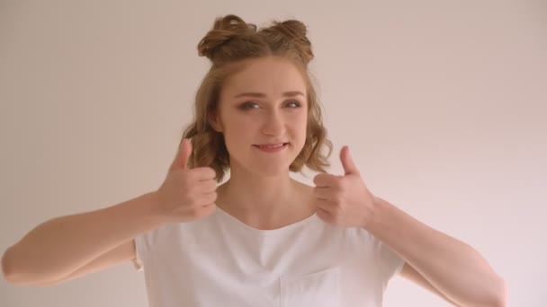 Closeup shoot of young cute caucasian female with hair buns showing thumbs up smiling happily looking at camera in the white room indoors — Stock Video