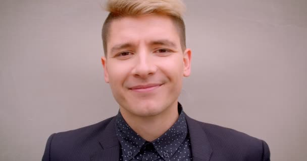 Closeup shoot of young attractive caucasian blonde man smiling happily looking at camera with the wall on the background — Stock Video