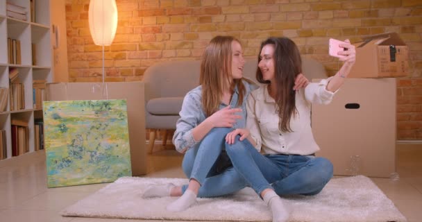 Closeup shoot of young beautiful lesbian couple talking on camera streaming live on the phone hugging happily and sitting on the floor in a newly bought apartment — Stock Video