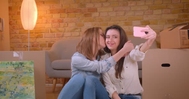 Closeup shoot of young beautiful lesbian couple taking selfies on the phone kissing happily and sitting on the floor in a newly bought apartment — Stock Video