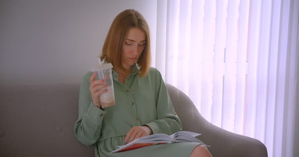 Retrato de cerca de una joven estudiante caucásica leyendo un libro sentado en un sofá con refresco en un paquete ecológico . — Vídeo de stock