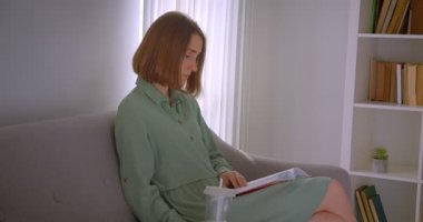 Oturma odasında kanepede oturan beyaz genç kız öğrenci okuma kitabının yakın profil portresi.