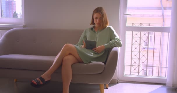 Retrato de una joven estudiante sexy trabajando con una tableta sentada en un sofá en la sala de estar . — Vídeos de Stock