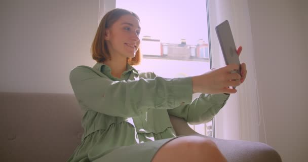 Retrato de una joven estudiante sexy haciendo selfies en una tableta sentada en un sofá en la sala de estar . — Vídeos de Stock