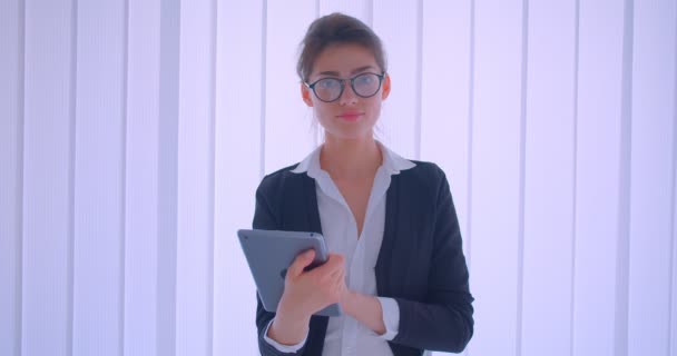 Primer plano de la joven mujer de negocios bastante caucásica sosteniendo una tableta y mirando a la cámara sonriendo felizmente en el interior de una habitación blanca — Vídeo de stock