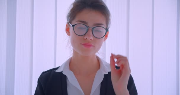 Primer plano de la joven mujer de negocios bastante caucásica y la fijación de sus gafas y mirando a la cámara sonriendo alegremente en el interior de una habitación blanca — Vídeo de stock