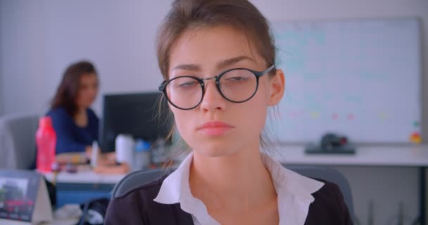 Nahaufnahme einer jungen kaukasischen Geschäftsfrau mit Brille, die im Büro in die Kamera schaut, im Hintergrund Mitarbeiter — Stockvideo