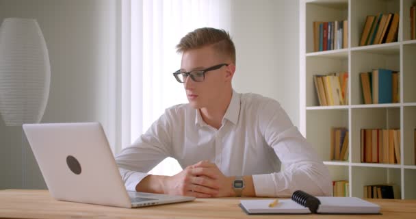 Close-up portret van jonge knappe Kaukasische zakenman in glazen met een video-oproep op de laptop binnenshuis in het kantoor — Stockvideo