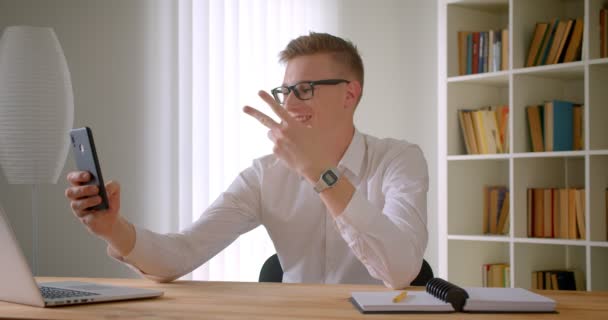 Closeup portrait of young handsome caucasian businessman in glasses having a video call on the phone talking cheerfully indoors in the office — Stock Video