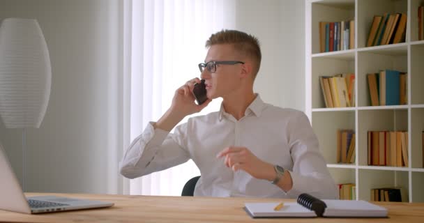 オフィスで屋内で電話を持つ眼鏡で若いハンサムな白人ビジネスマンのクローズアップ肖像画 — ストック動画