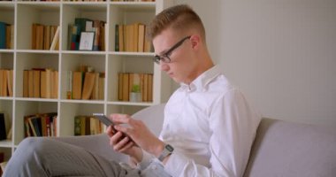 Genç yakışıklı beyaz işadamının gözlüklü yakın çekim portresi tableti kullanarak kameraya bakarak kanepede otururken içeride mutlu bir şekilde oturuyor