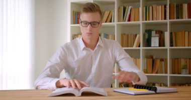 Üniversite kütüphanesinde kameraya bakarak masanın yanında oturan bir kitap okuyan genç yakışıklı beyaz öğrencinin yakın çekim portresi