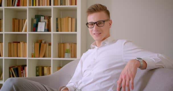 Closeup portræt af ung flot kaukasisk forretningsmand i briller smilende glad sidder på sofaen ser på kameraet indendørs – Stock-video