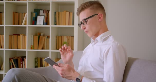 Nahaufnahme Porträt eines jungen gutaussehenden kaukasischen Geschäftsmannes mit Brille, der das Tablet benutzt und in die Kamera schaut, die drinnen auf der Couch sitzt — Stockvideo